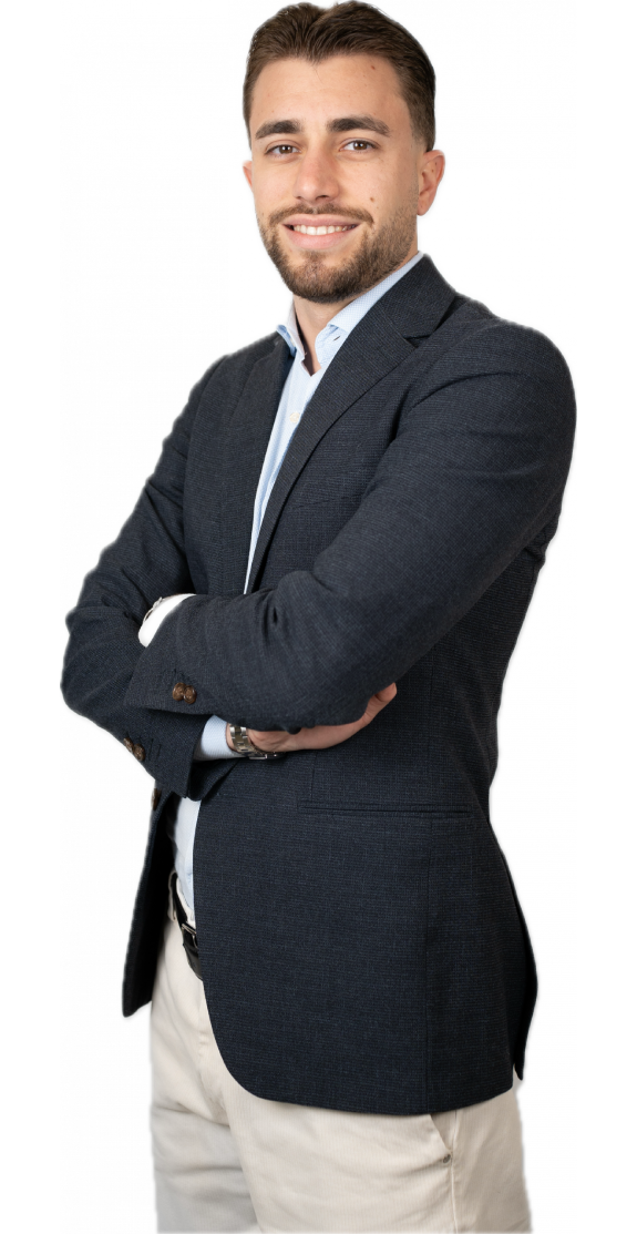 Un giovane uomo con capelli corti e barba curata sta in piedi con sicurezza a braccia incrociate. Indossa un blazer blu, una camicia azzurra e pantaloni beige e sorride all'obiettivo su uno sfondo bianco.
