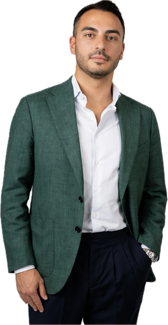 Un uomo in piedi su un semplice sfondo bianco indossa un blazer verde su una camicia bianca e pantaloni scuri. Ha i capelli corti e scuri, la barba ben curata e un'espressione neutra, con una mano in tasca e l'altra appoggiata al fianco.